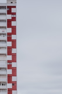 A multi-story apartment building with a distinctive red and white checkered facade. The structure is positioned on the left side of the image, leaving a large expanse of overcast sky to the right. The building features balconies, windows, and a sign indicating something is for rent or sale.