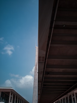 A modern architectural scene showcasing a tall building with a spire and an adjacent overpass or covered walkway. The structure's design features sleek lines and large glass windows. A portion of the sky is visible, displaying a few clouds against a blue backdrop.