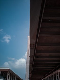 A modern architectural scene showcasing a tall building with a spire and an adjacent overpass or covered walkway. The structure's design features sleek lines and large glass windows. A portion of the sky is visible, displaying a few clouds against a blue backdrop.