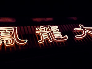 Neon Chinese characters glow brightly in the dark, mounted against a slatted background. The lights exhibit a warm, inviting hue with a luminous intensity.