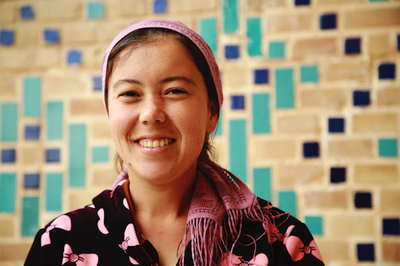 Aya El Jazzar smiling warmly while speaking in front of a camera, with a cozy, inviting background.