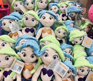 A collection of plush mermaid dolls with vibrant blue and green hair. Each doll features colorful shell tops, either in orange or purple. They have cheerful expressions and are densely packed together with some tags visible.