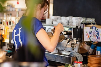 A person wearing a blue shirt with the letter 'H' on the back is preparing a drink using a professional espresso machine. Several white coffee cups are stacked on top of the machine. The setting appears to be a cozy café with various bottles and decorations adorning the background.