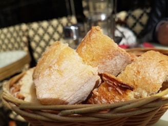 A close-up view of a basket filled with crispy, golden-brown bread pieces. The bread is beautifully textured, with a slightly crispy crust and a soft interior. The basket is wicker, providing a rustic appearance, and is placed on an outdoor table with a blurred background, suggesting a café setting.