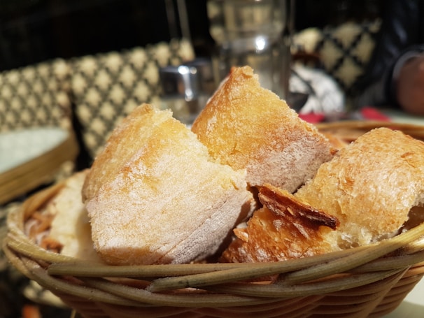 A close-up view of a basket filled with crispy, golden-brown bread pieces. The bread is beautifully textured, with a slightly crispy crust and a soft interior. The basket is wicker, providing a rustic appearance, and is placed on an outdoor table with a blurred background, suggesting a café setting.