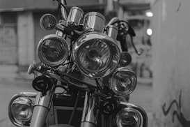 A close-up view of a motorcycle's front, featuring multiple round headlights and intricate details of the metal components. The image is in black and white, highlighting the reflective surfaces and textures of the motorcycle parts.