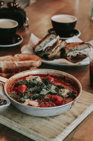 A hearty meal consisting of a bowl of tomato-based stew garnished with fresh herbs and red peppers, accompanied by toasted bread. In the background, there are sandwiches filled with various ingredients, and cups of coffee or tea are placed alongside.