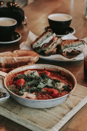 A hearty meal consisting of a bowl of tomato-based stew garnished with fresh herbs and red peppers, accompanied by toasted bread. In the background, there are sandwiches filled with various ingredients, and cups of coffee or tea are placed alongside.