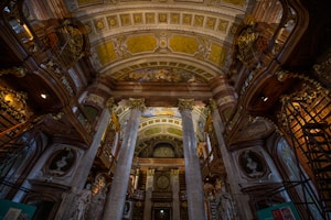 An ornate library interior features grand columns, intricate ceiling paintings, and tall, elaborately carved wooden bookshelves. The architecture boasts a Baroque style with detailed frescoes and gold embellishments. Soft lighting adds a warm ambiance to the scene.