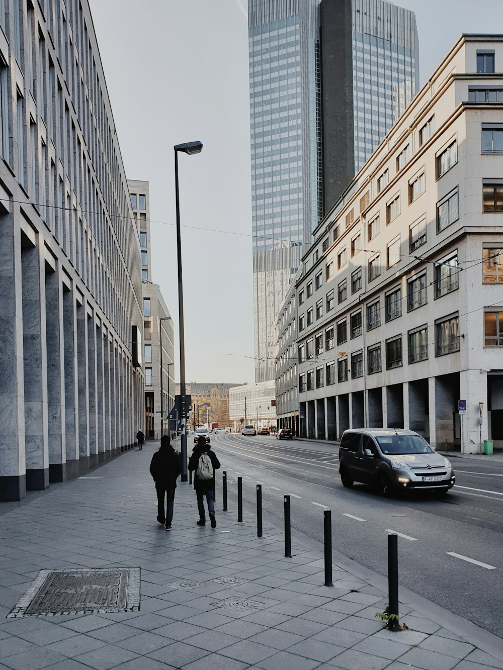 dos personas caminando cerca de la carretera y un SUV gris en la carretera al lado de edificios de gran altura