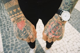 A close-up of two tattooed hands, showcasing intricate designs including roses and geometric patterns. The person is wearing a wristwatch on the right hand, and the background consists of a tiled stone floor.