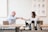 Two people are sitting across from each other in a modern cafe, shaking hands and smiling. A large poster on the wall behind them features various botanical illustrations. The counter in front of them holds glasses, coffee pots, and other cafe items. Each person has a laptop in front of them.