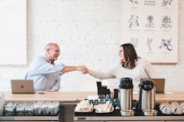 Two people are sitting across from each other in a modern cafe, shaking hands and smiling. A large poster on the wall behind them features various botanical illustrations. The counter in front of them holds glasses, coffee pots, and other cafe items. Each person has a laptop in front of them.