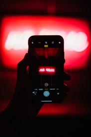 A hand holding a smartphone with the camera app open, capturing a bright neon red sign that is blurred in the background. The phone displays a clearer view of the sign, which has text written in Russian.