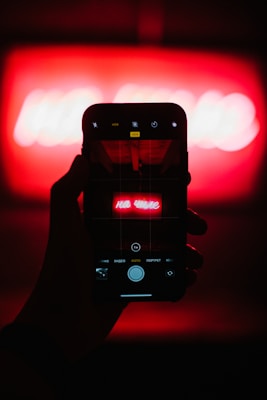 A hand holding a smartphone with the camera app open, capturing a bright neon red sign that is blurred in the background. The phone displays a clearer view of the sign, which has text written in Russian.