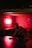 A man playing table tennis in a dimly lit room with red lighting. The walls are covered with graffiti-style writing, and there's a small picture frame illuminated on one wall. The mood is casual and relaxed.