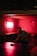 A man playing table tennis in a dimly lit room with red lighting. The walls are covered with graffiti-style writing, and there's a small picture frame illuminated on one wall. The mood is casual and relaxed.