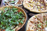 A variety of pizza dishes are placed on stands, each with different toppings. One pizza is topped with fresh arugula and a drizzle of balsamic glaze, another features a mix of diced mango and onions, and a third is decorated with green onions and melted cheese. In the background, there are additional dishes including a salad and some appetizer snacks.