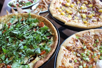 A variety of pizza dishes are placed on stands, each with different toppings. One pizza is topped with fresh arugula and a drizzle of balsamic glaze, another features a mix of diced mango and onions, and a third is decorated with green onions and melted cheese. In the background, there are additional dishes including a salad and some appetizer snacks.