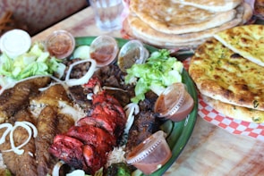 A large platter filled with a variety of grilled meats including a red tandoori dish and other brown grilled items accompanied by white onions and leafy greens. Small containers of various sauces encircle the platter, which sits on a table next to stacks of naan bread. The background shows a glass of water.