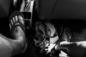 Black and white photo depicting the interior of a vehicle focusing on the floor area. Two bare feet wearing sandals are visible, along with another pair of sandals and clutter, such as straps or belts, near the center console.