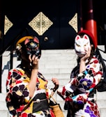 Two individuals dressed in colorful kimonos stand on steps holding fox masks to their faces. The kimonos feature vibrant floral patterns with shades of red, yellow, and blue. Behind them, decorative, ornate panels adorn a dark wall, adding to the cultural and traditional theme of the image. A basket and small handbags complement their attire.