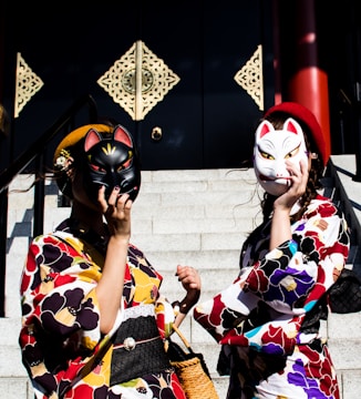 Two individuals dressed in colorful kimonos stand on steps holding fox masks to their faces. The kimonos feature vibrant floral patterns with shades of red, yellow, and blue. Behind them, decorative, ornate panels adorn a dark wall, adding to the cultural and traditional theme of the image. A basket and small handbags complement their attire.