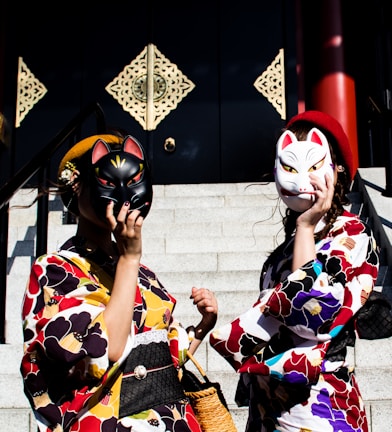 Two individuals dressed in colorful kimonos stand on steps holding fox masks to their faces. The kimonos feature vibrant floral patterns with shades of red, yellow, and blue. Behind them, decorative, ornate panels adorn a dark wall, adding to the cultural and traditional theme of the image. A basket and small handbags complement their attire.