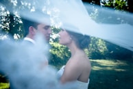 A romantic moment between a couple standing closely in a tranquil outdoor setting, partially obscured by a flowing white veil. The background is filled with lush green foliage and soft, diffused sunlight.