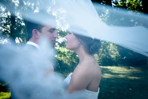 A romantic moment between a couple standing closely in a tranquil outdoor setting, partially obscured by a flowing white veil. The background is filled with lush green foliage and soft, diffused sunlight.