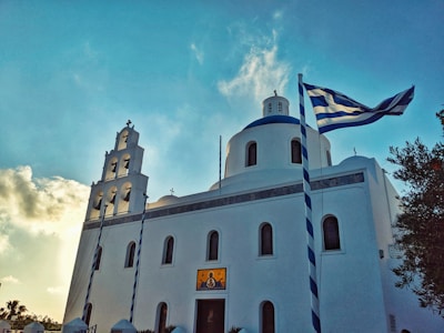 A white church with a blue dome is surrounded by clear blue skies. The architecture features a bell tower with multiple arches and Greek flags waving in the wind. The sun creates a warm glow as it sets or rises, casting soft shadows.