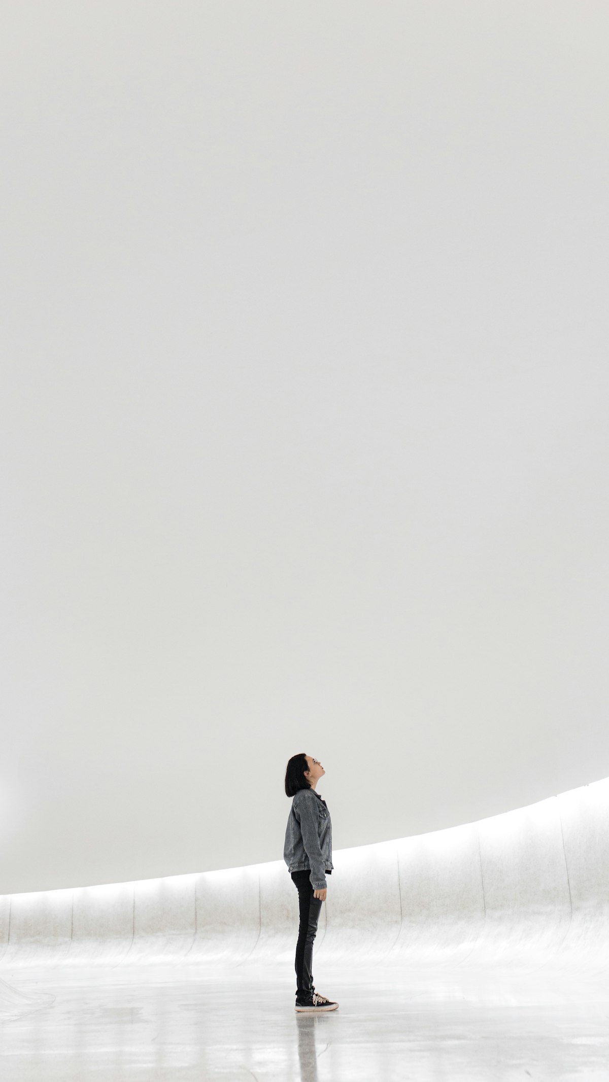A lone figure standing in a vast luminous white space gazing upward, scale-and-diminishing-returns register