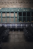 Rows of empty metal and black leather chairs are arranged in a waiting area with a vintage sign above reading 'Quebec Central Railway Canadian National Railway.' The wall behind features green painted paneling and brickwork, creating a nostalgic and historic atmosphere.