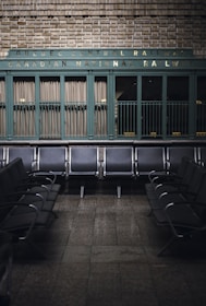 Rows of empty metal and black leather chairs are arranged in a waiting area with a vintage sign above reading 'Quebec Central Railway Canadian National Railway.' The wall behind features green painted paneling and brickwork, creating a nostalgic and historic atmosphere.
