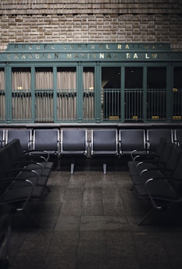 Rows of empty metal and black leather chairs are arranged in a waiting area with a vintage sign above reading 'Quebec Central Railway Canadian National Railway.' The wall behind features green painted paneling and brickwork, creating a nostalgic and historic atmosphere.