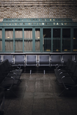 Rows of empty metal and black leather chairs are arranged in a waiting area with a vintage sign above reading 'Quebec Central Railway Canadian National Railway.' The wall behind features green painted paneling and brickwork, creating a nostalgic and historic atmosphere.