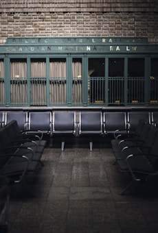 Rows of empty metal and black leather chairs are arranged in a waiting area with a vintage sign above reading 'Quebec Central Railway Canadian National Railway.' The wall behind features green painted paneling and brickwork, creating a nostalgic and historic atmosphere.