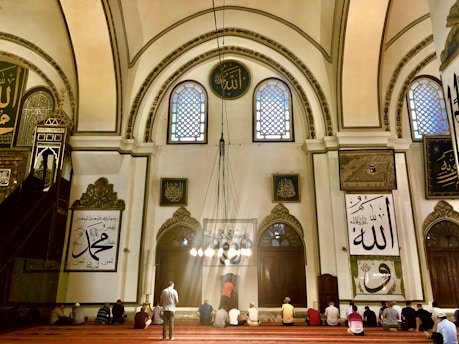 A group of people sits on a red-carpeted floor within a grand, ornately decorated mosque. The architecture features high arched ceilings with intricate patterns and calligraphy. Large windows allow light to stream through, illuminating the interior with a warm glow. Religious inscriptions and artwork are present on the walls, adding to the sacred atmosphere.