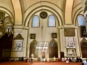 A group of people sits on a red-carpeted floor within a grand, ornately decorated mosque. The architecture features high arched ceilings with intricate patterns and calligraphy. Large windows allow light to stream through, illuminating the interior with a warm glow. Religious inscriptions and artwork are present on the walls, adding to the sacred atmosphere.