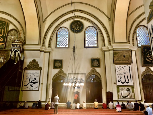 A group of people sits on a red-carpeted floor within a grand, ornately decorated mosque. The architecture features high arched ceilings with intricate patterns and calligraphy. Large windows allow light to stream through, illuminating the interior with a warm glow. Religious inscriptions and artwork are present on the walls, adding to the sacred atmosphere.
