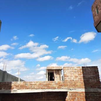 A partly constructed brick building extends into a bright blue sky with scattered white clouds. Unfinished concrete and exposed rebar indicate ongoing construction work.
