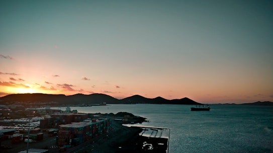 A serene waterfront scene during sunset with a container port in the foreground and a large ship on the calm water. The sky is a gradient of warm oranges and cool blues as the sun sets behind distant hills. Shipping containers are stacked near the water, and the entire area has a tranquil and peaceful atmosphere.