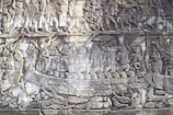 Intricately carved stone relief depicting a historical battle scene. Multiple figures are shown engaging in combat, wielding weapons, and riding chariots. The figures are dressed in ancient attire, suggesting a setting from a bygone era.