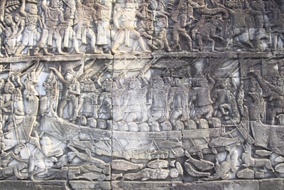 Intricately carved stone relief depicting a historical battle scene. Multiple figures are shown engaging in combat, wielding weapons, and riding chariots. The figures are dressed in ancient attire, suggesting a setting from a bygone era.