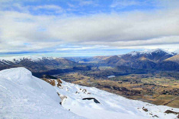 View from a snowy Coronet Peak ski slope over the Wakatipu basin toward Arrowtown, with snow-capped ranges on the horizon