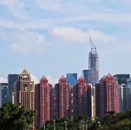 A cityscape featuring a series of tall, red residential buildings with pyramid-shaped roofs, set against a backdrop of modern skyscrapers. A construction crane is visible atop one of the buildings, indicating ongoing development. The foreground includes green trees, and the sky is mostly clear with some scattered clouds.