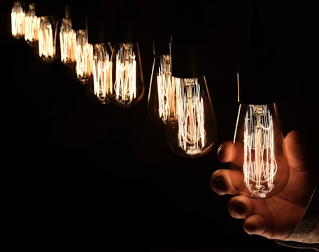 A series of vintage-style filament light bulbs are illuminated against a dark background. The bulbs emit a warm glow, and one of them is being held by a hand. The focus is on the intricate filaments inside the bulbs, creating a cozy and nostalgic atmosphere.