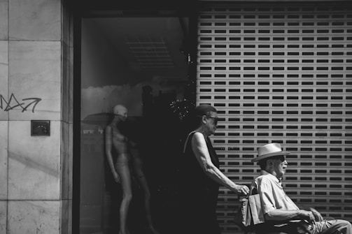 A black and white photograph depicts an elderly man sitting in a wheelchair, being pushed by a woman. They are in front of a building with a large reflective glass window, which holds a mannequin inside. The shuttered background adds to the urban setting, and some graffiti is visible on the left wall.