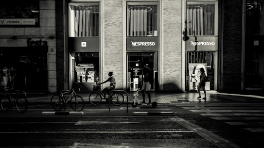 A monochromatic street scene featuring people walking and cycling in front of a Nespresso store. The setting is urban, with a focus on architecture and the interactions of pedestrians. Shadows and light create a dramatic contrast, evoking a sense of tranquility and contemplation.