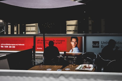 The image depicts a modern indoor setting with posters on a wall featuring a professional headshot and text related to marketing. Silhouetted figures are seated at tables in the foreground, casting shadows. The lighting is dim, creating contrast between the illuminated posters and darker seating area.